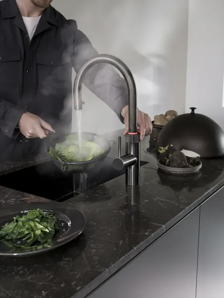 Man steaming leafy greens in a pan under running water from a modern kitchen faucet