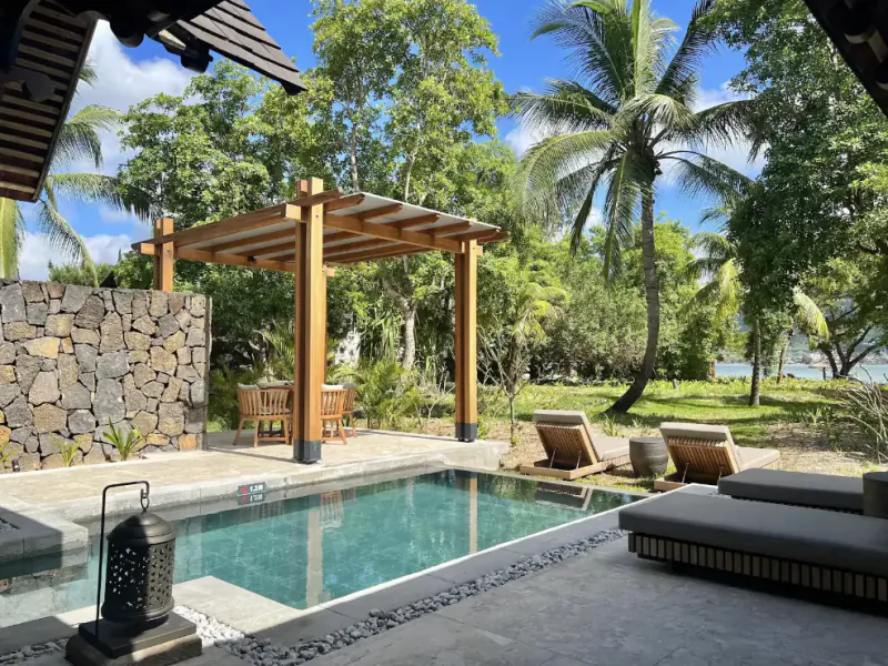 Luxury resort pool with wooden pergola, lounge chairs, stone wall, and tropical palm trees overlooking the ocean
