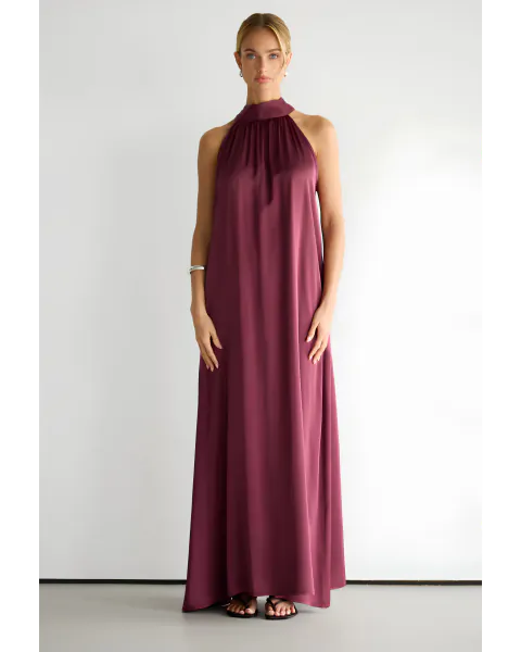 Woman in a long burgundy satin halter-neck dress standing against a white studio backdrop