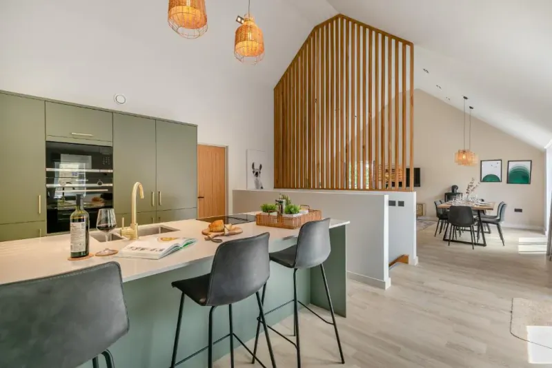 Modern open-plan kitchen and dining area with sage green cabinets, white island, grey bar stools, and wooden room divider