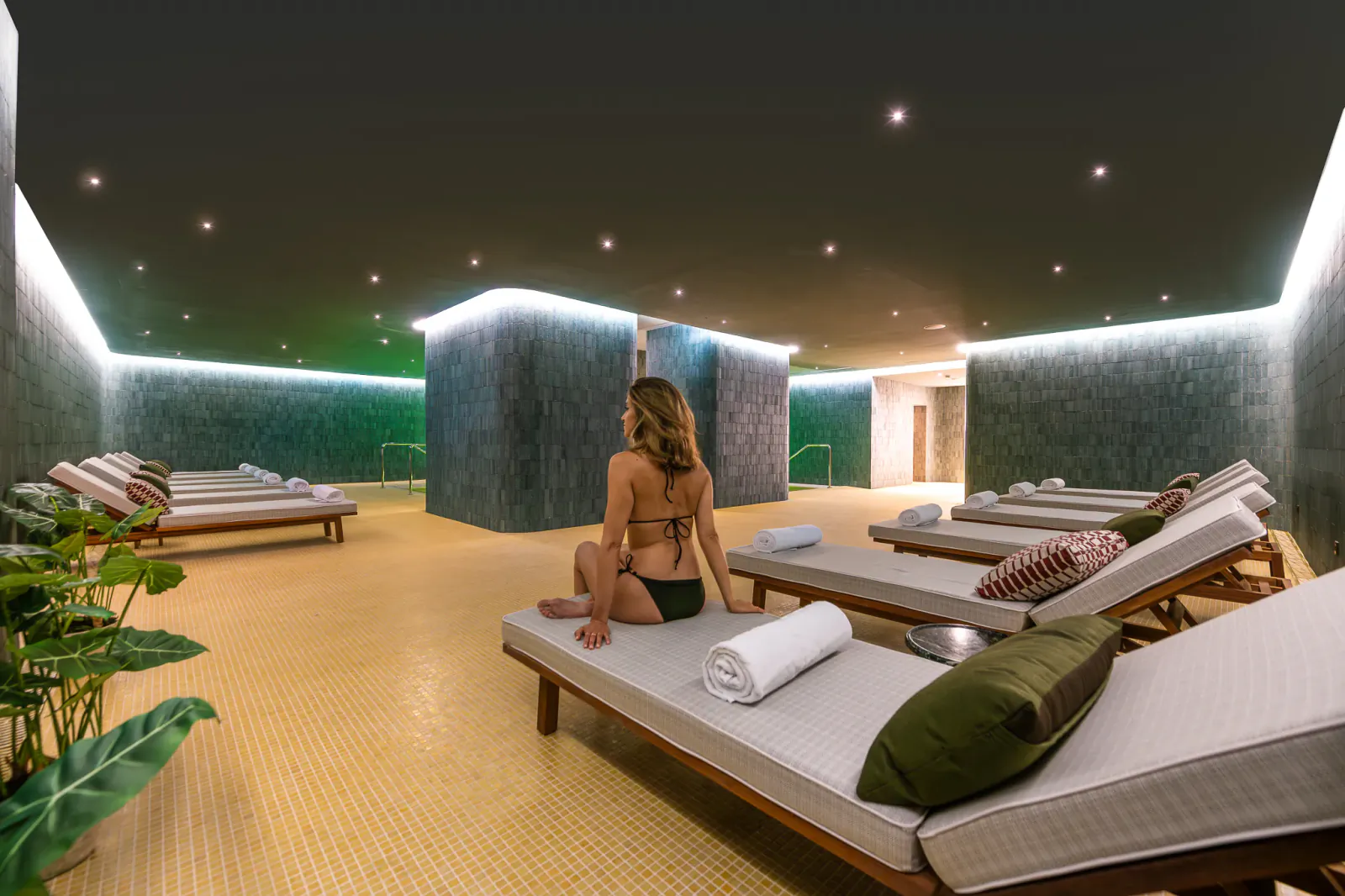 Woman in black bikini sitting on lounge chair in modern spa with starlit ceiling and tiled walls