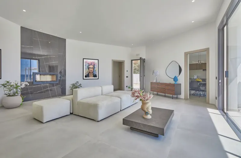 Modern open-plan living room with white sectional sofa, stone coffee table, and large glass doors opening to a bright interior