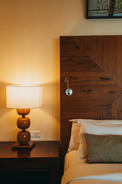 Bedside table with lit wooden lamp beside a bed with white pillows and brown headboard