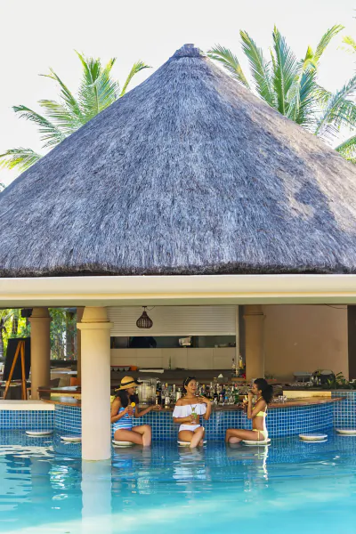 Three women sitting at a swim-up bar under a thatched roof at a tropical resort pool with palm trees
