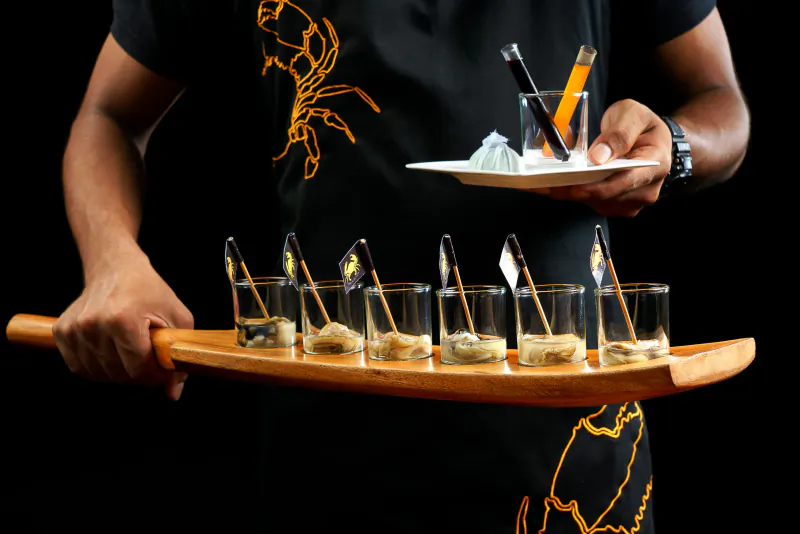 Person holding a wooden tray with six small glasses of food and a white plate with three test tubes of colorful liquids