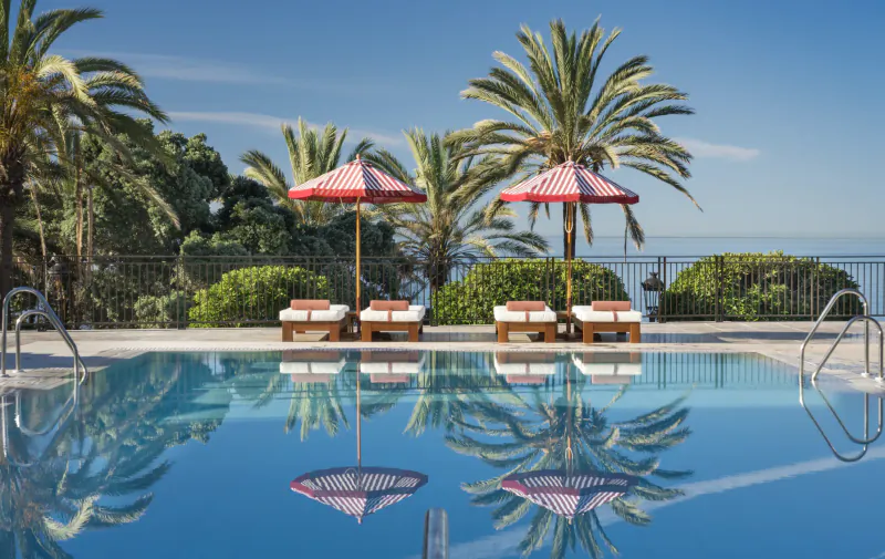 Luxury poolside with red striped umbrellas, lounge chairs, palm trees, and calm blue water reflecting the sky