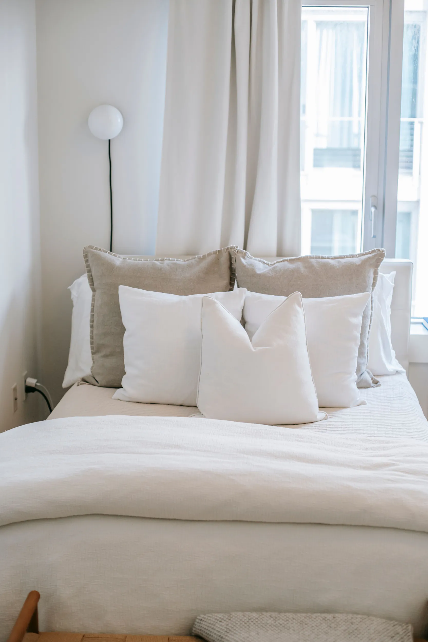 Minimalist bedroom with neatly made bed featuring white and beige pillows against a white wall, floor lamp, and window with sheer curtains