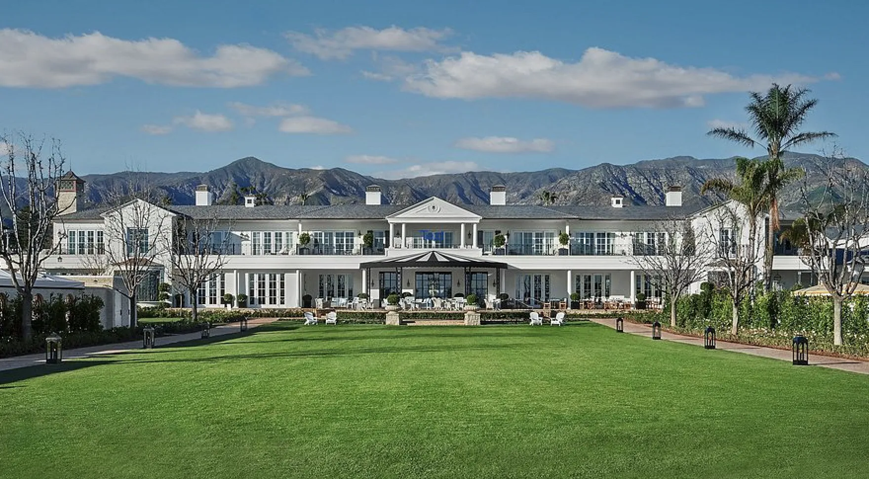 Large white mansion with lawn, patio furniture, and mountain backdrop