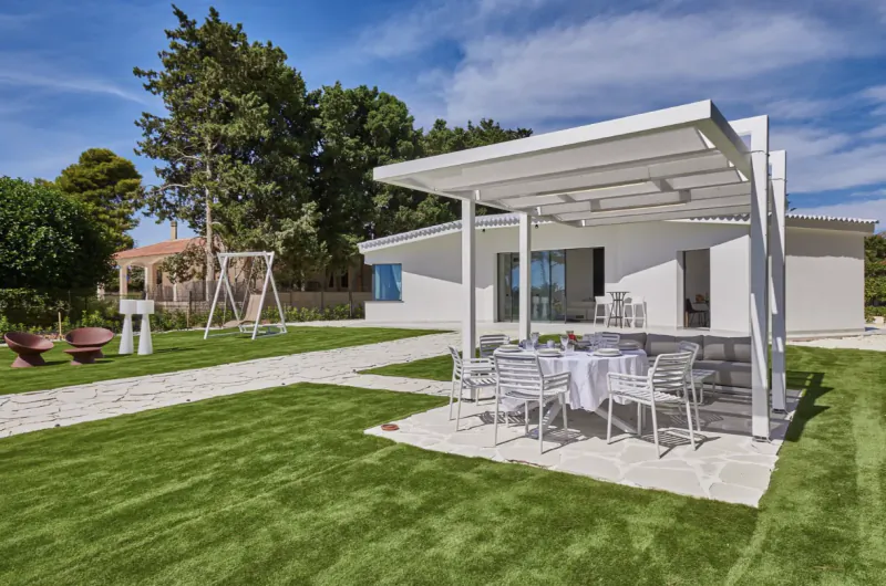 Modern white villa with covered patio, dining table, and chairs on green lawn with swing set