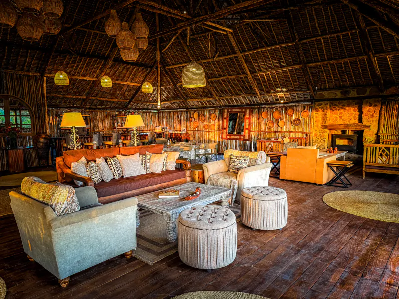 Spacious safari lodge lounge with wooden sofas, tufted ottomans, woven lamps, and a fireplace under a thatched ceiling