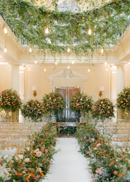 Elegant wedding aisle lined with lush peach and orange floral arrangements leading to ornate double doors under a canopy of hanging greenery and glowing bulbs