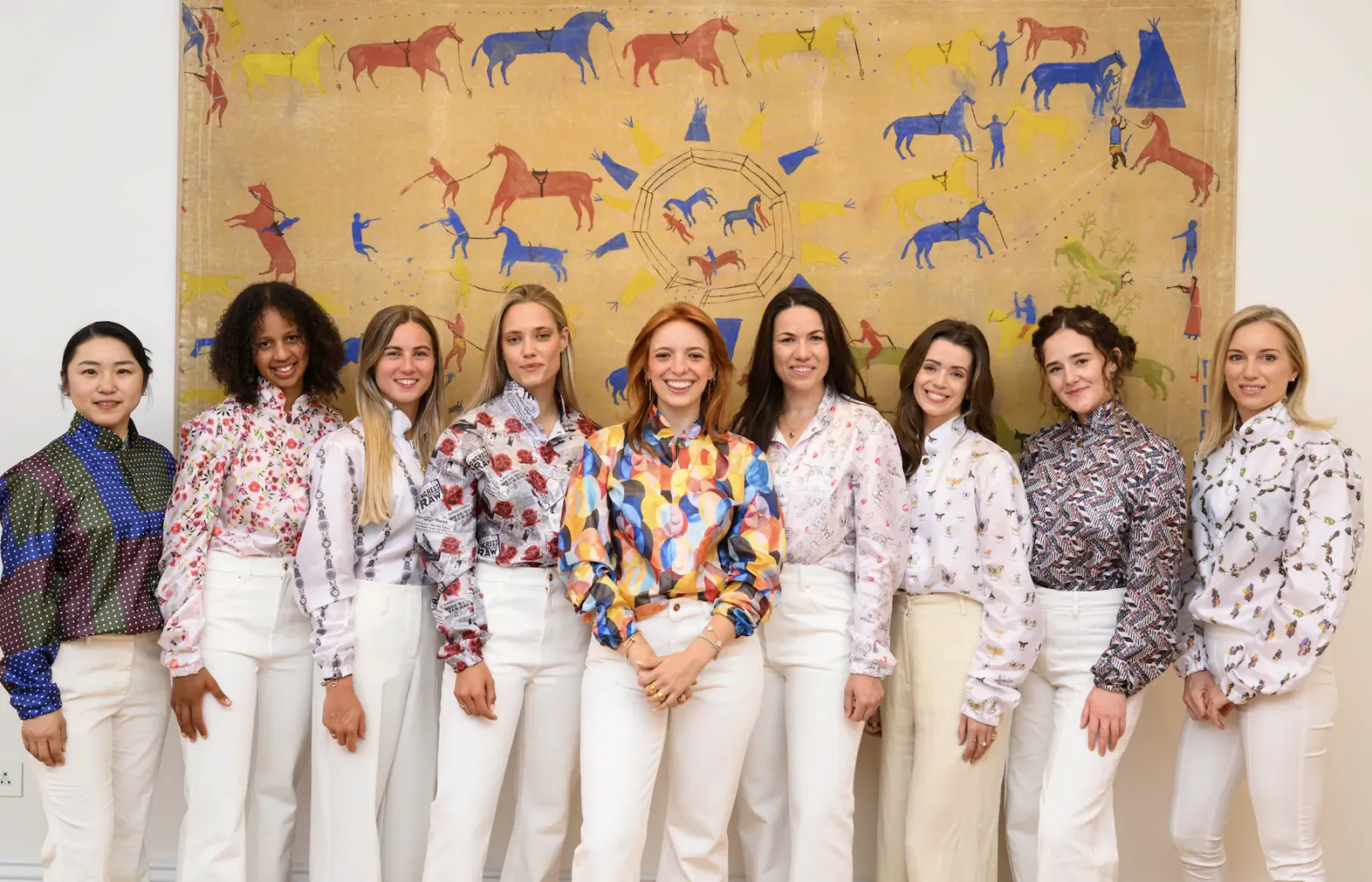 Nine women in white trousers and patterned blouses pose smiling in front of a large colorful horse mural
