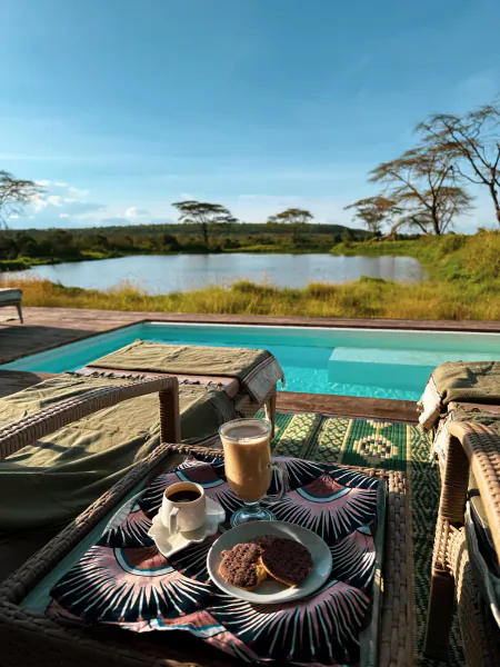 Breakfast tray with coffee, latte and cookies on a patterned cloth beside a pool overlooking a savanna lake