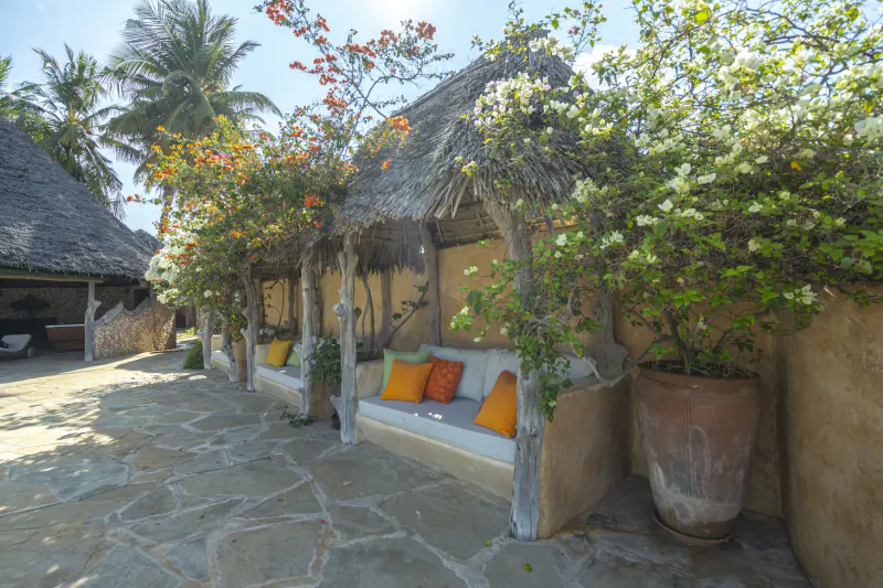 Outdoor seating area at Manda Bay Lodge with thatched-roof bench, colorful cushions, flowering trees, and stone patio