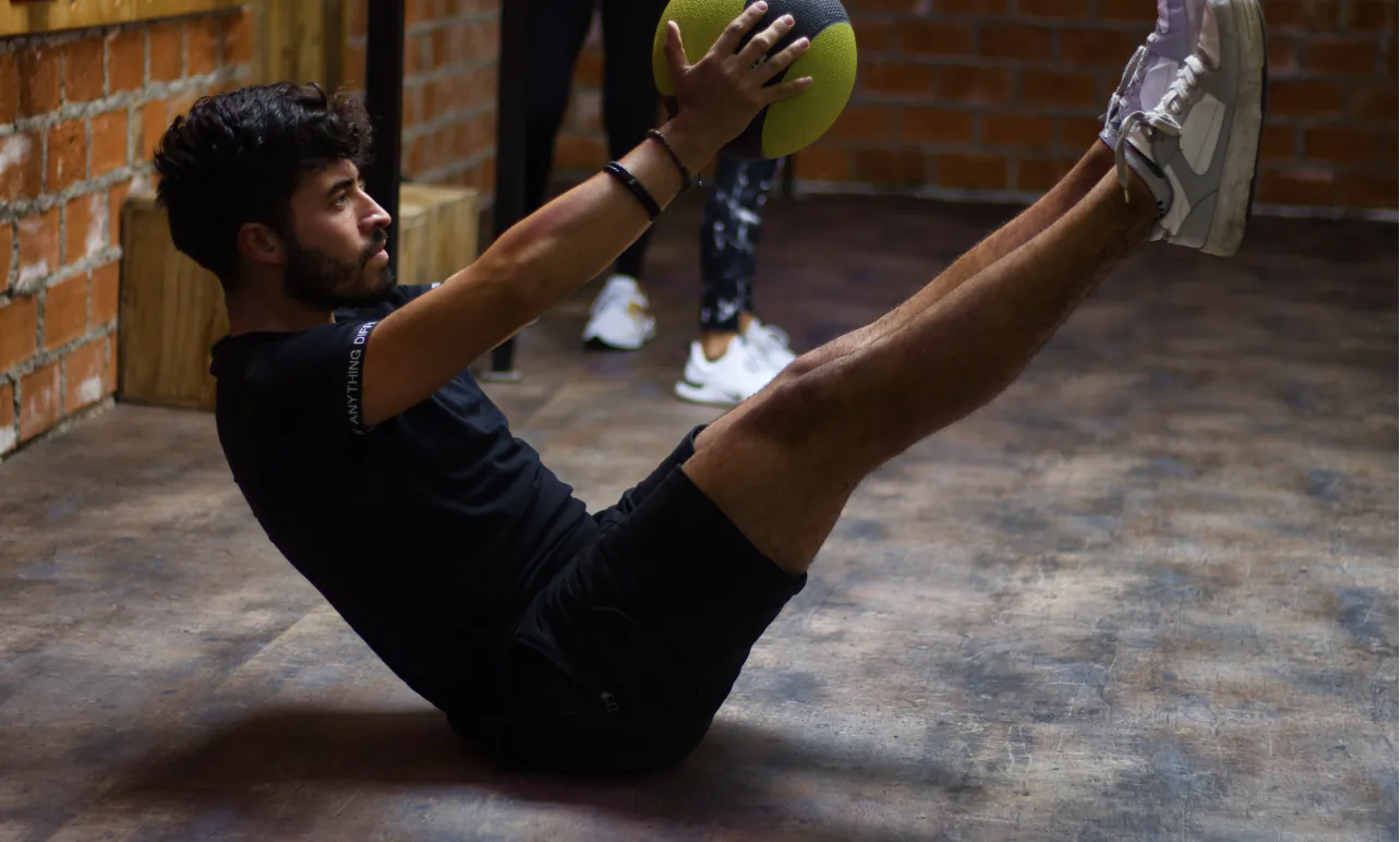Man in black workout clothes holding a green medicine ball while performing a V-sit exercise on a gym floor