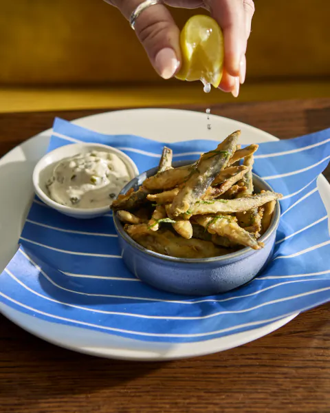 Hand squeezing lemon over a bowl of fried fish with dipping sauce on a blue striped napkin