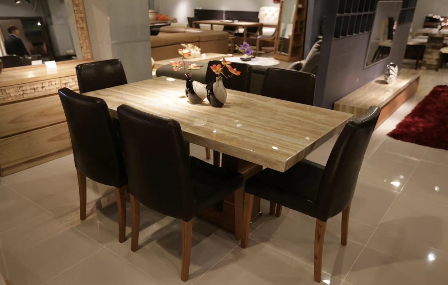 Modern dining room with light wood table, four black leather chairs, and decorative vases with flowers in a furniture showroom
