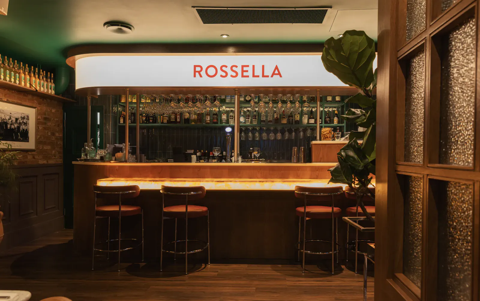 Rossella bar with glowing counter, three bar stools, and backlit shelves of bottles
