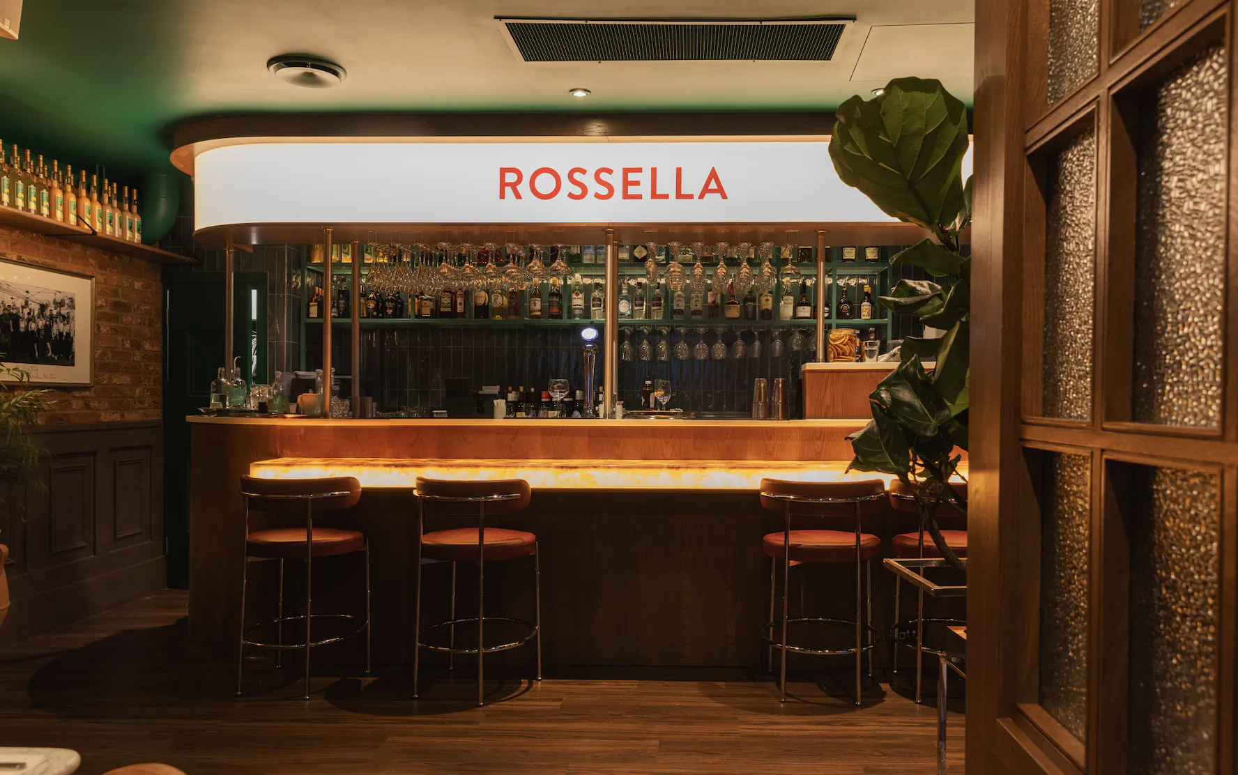 Rossella bar with glowing counter, three bar stools, and backlit shelves of bottles