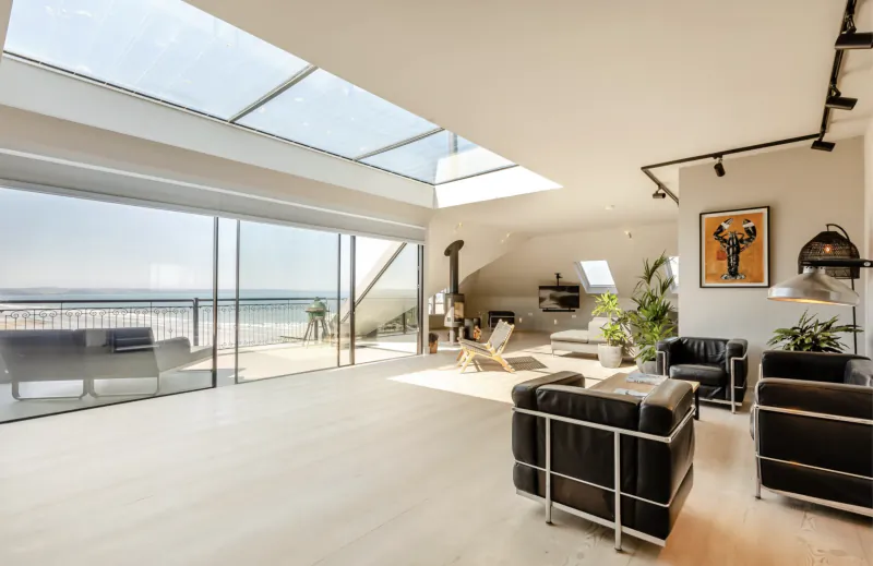 Modern open-plan luxury living room with black leather armchairs, skylights, and floor-to-ceiling glass doors opening to a sea-view balcony