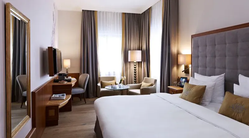 Modern hotel room with large bed, tufted headboard, seating area, desk, TV, and window with curtains