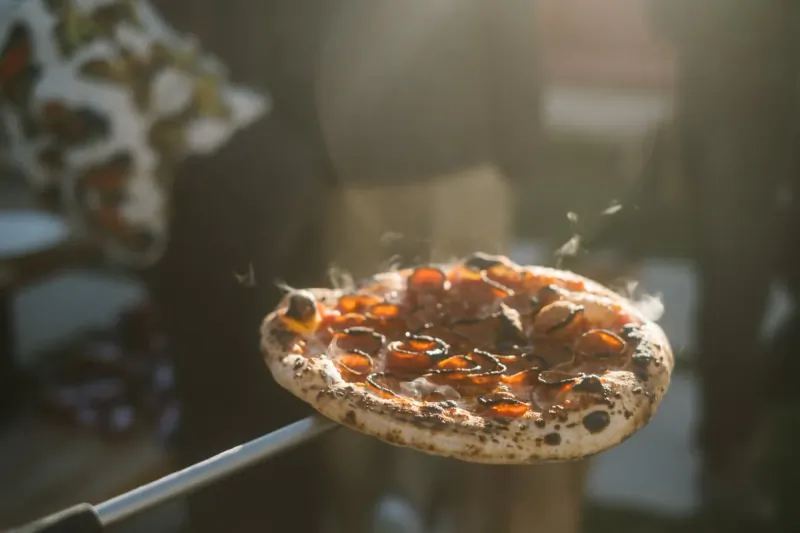 Pepperoni pizza on a peel with steam rising, held outdoors in sunlight