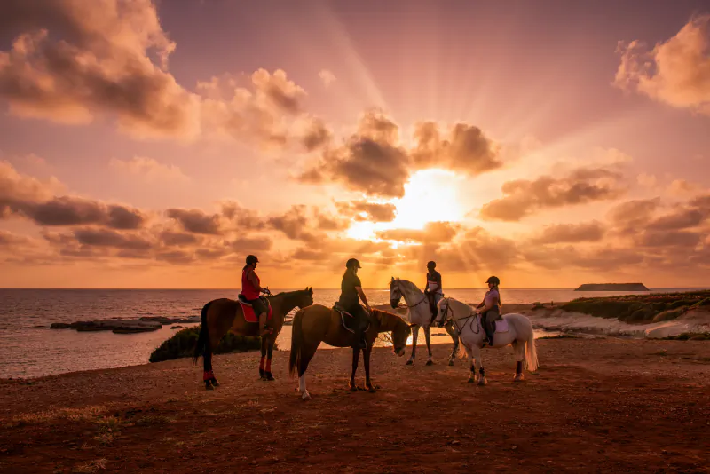 Four riders on horseback silhouetted against a dramatic sunset over the sea at Cap St Georges, Cyprus