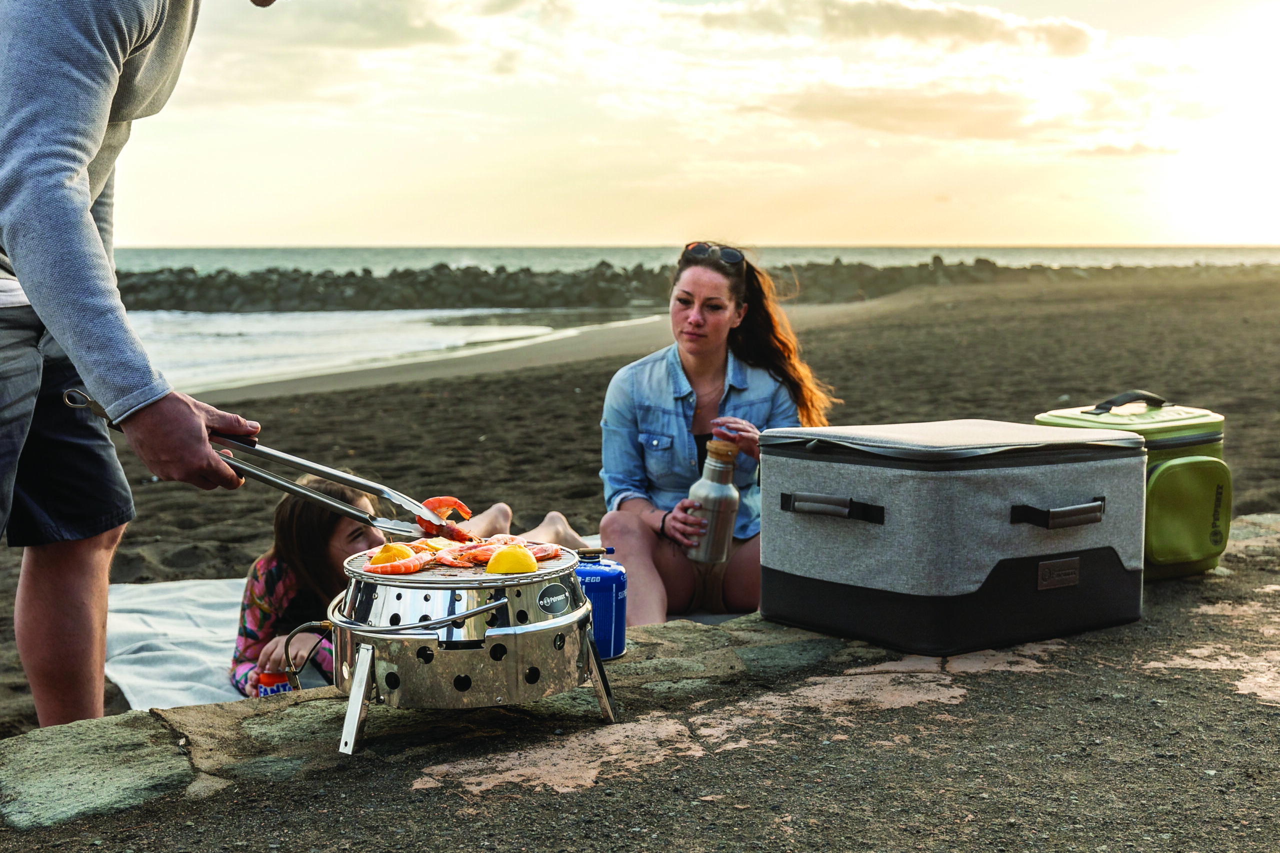Family grilling hot dogs on a portable fire pit at sunset on a sandy beach with coolers nearby