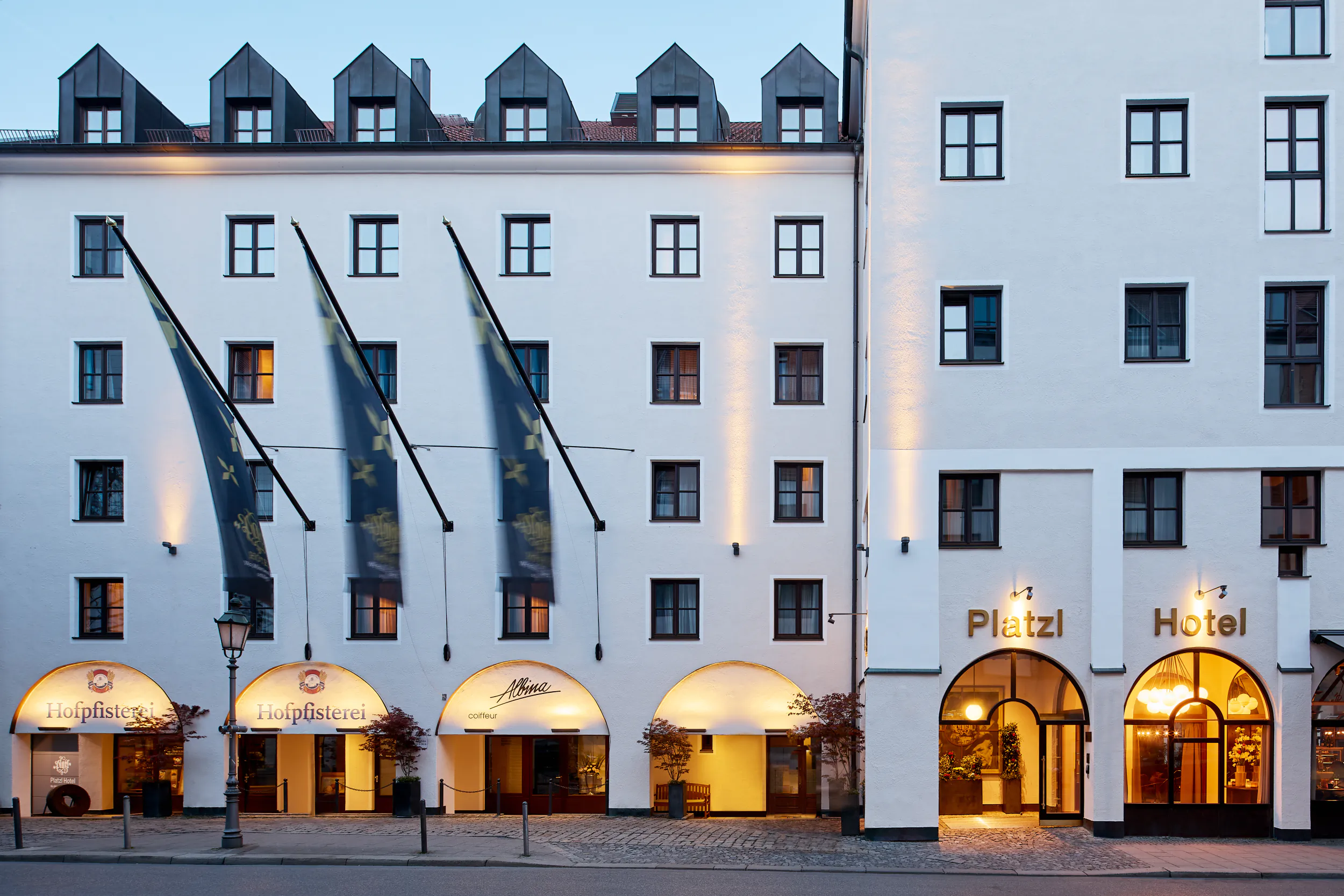 Platzl Hotel Munich facade at dusk with three flags, arched entrances, and illuminated signage