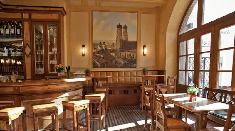 Elegant wooden bar and dining area with large cityscape painting, arched windows, and warm lighting in a Munich hotel