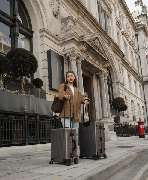 Woman in tan blazer standing with two suitcases outside a historic Post Office building