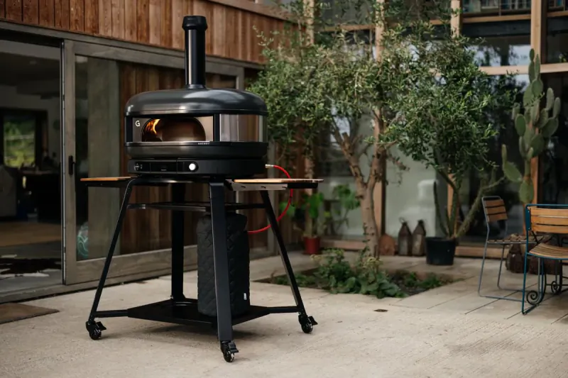 Gozney pizza oven on black stand with fire visible inside, outdoor patio setting
