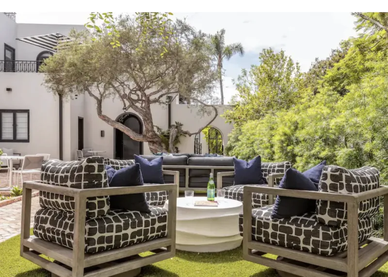 Modern outdoor patio with patterned armchairs, round coffee table, and dark sectional seating under a large tree in a lush garden