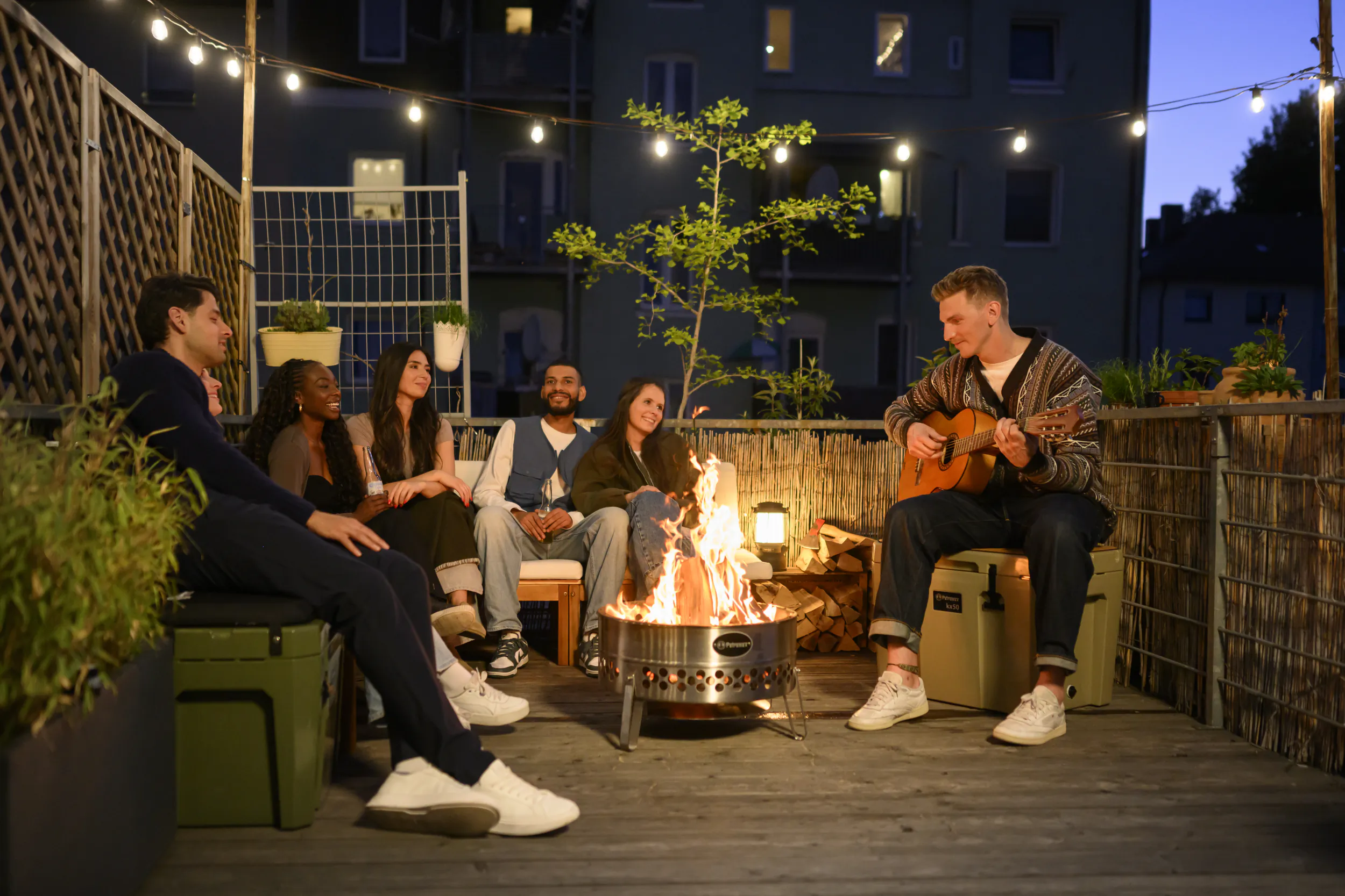 Friends gathered around a fire pit on a rooftop deck at dusk, one man playing guitar