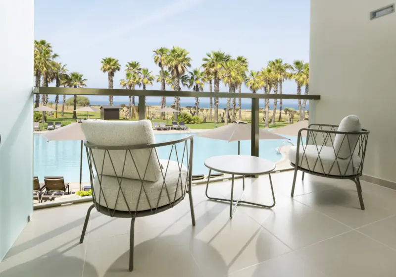 Balcony with two modern lounge chairs and a round table overlooking a pool, palm trees, and ocean at The Westin Salgados Beach Resort, Algarve