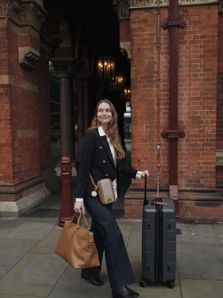 Woman in black blazer and wide-leg trousers smiling while holding a brown tote and pulling a grey suitcase outside a red brick building