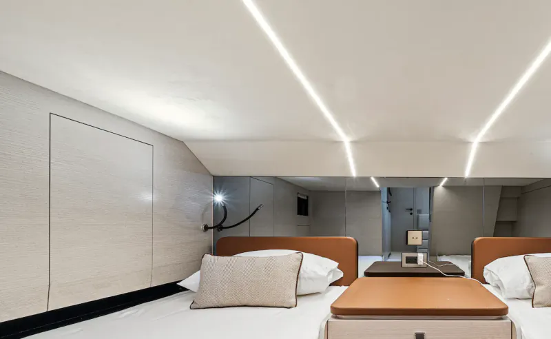 Modern yacht cabin with two beds, orange headboards, white linens, and sleek white ceiling with linear lighting