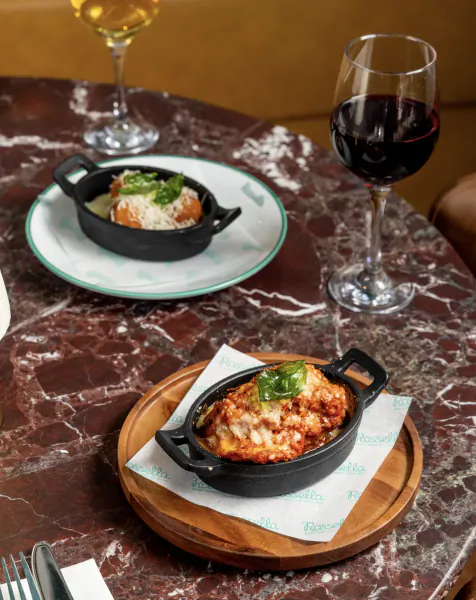 Two baked dishes in black skillets on a marble table with red wine and white wine glasses at Rossella restaurant