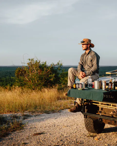 Man in safari hat and khaki shirt sits on a Toyota truck tailgate overlooking the African bush at sunset