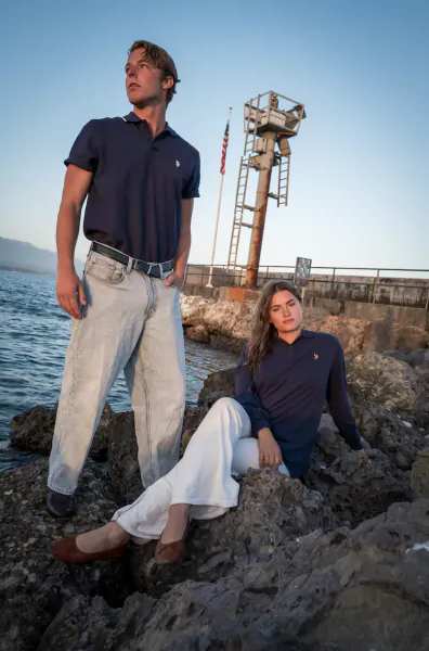 Young man and woman in navy polo shirts posing on rocky shore by water with a tall metal tower and American flag behind them