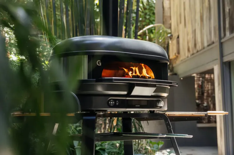 Black Gozney pizza oven with open flame inside on a stand, surrounded by plants and wooden deck