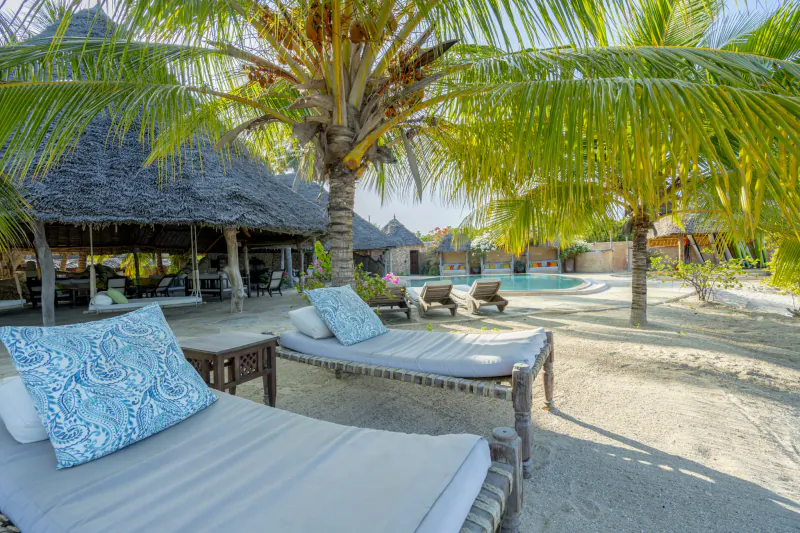 Beach lounge chairs with blue patterned pillows under palm trees at a tropical resort with thatched huts and pool