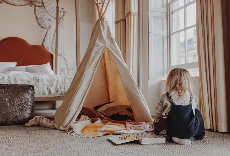 Toddler in overalls sits reading books inside a teepee tent in a cozy bedroom with a bed and large window