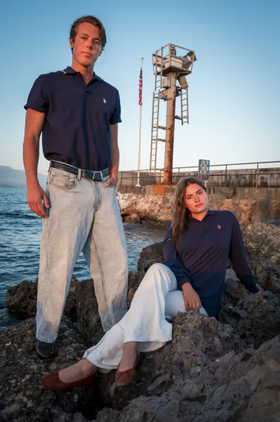 Young man in navy polo and light jeans standing beside woman in navy top and white pants seated on rocky shore with lifeguard tower and American flag behind them