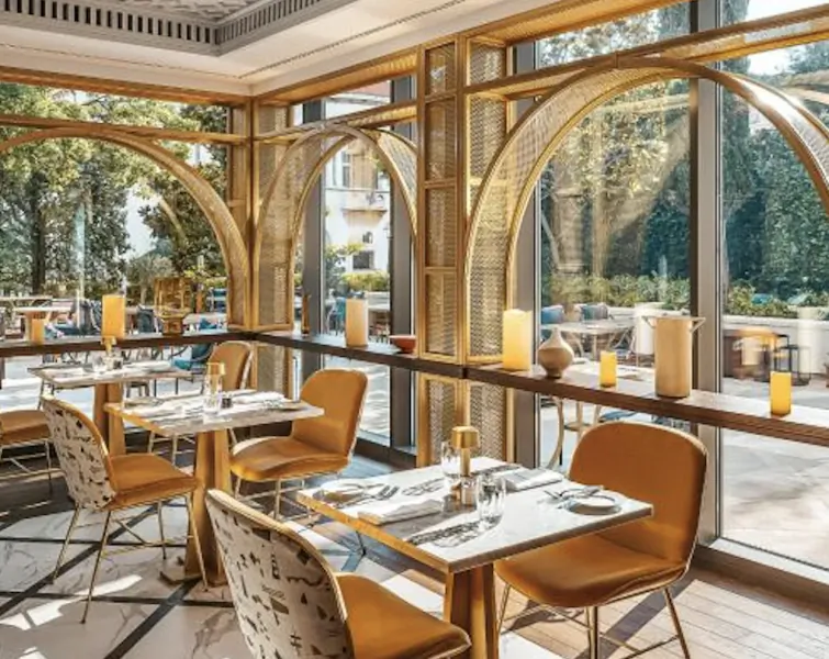 Elegant hotel restaurant with marble tables, mustard velvet chairs, and large arched windows overlooking a garden terrace