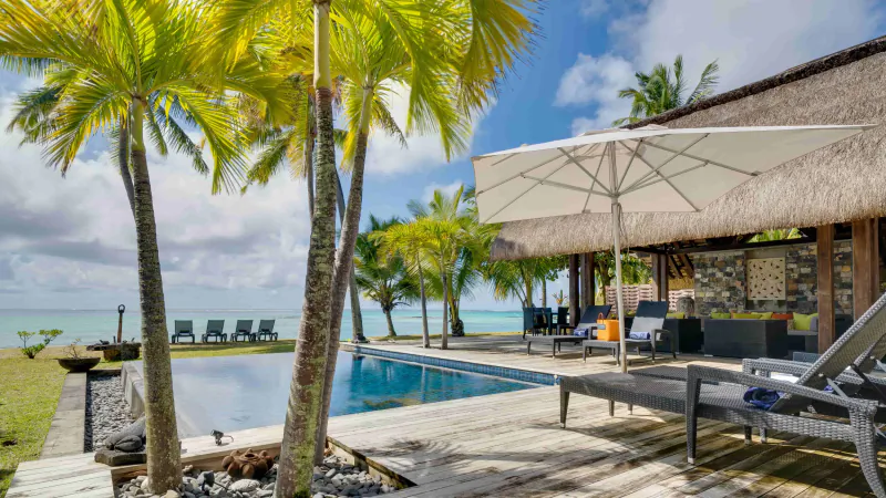 Luxury villa pool deck with lounge chairs, large umbrella, palm trees, and ocean view in Mauritius