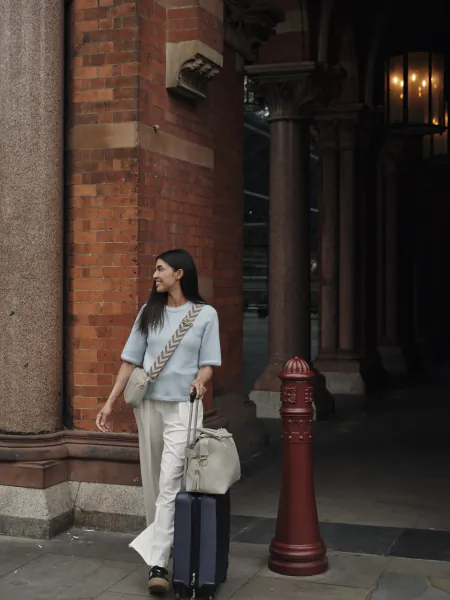 Woman with long dark hair in light blue top and white trousers pulling a navy suitcase outside a red brick building with columns