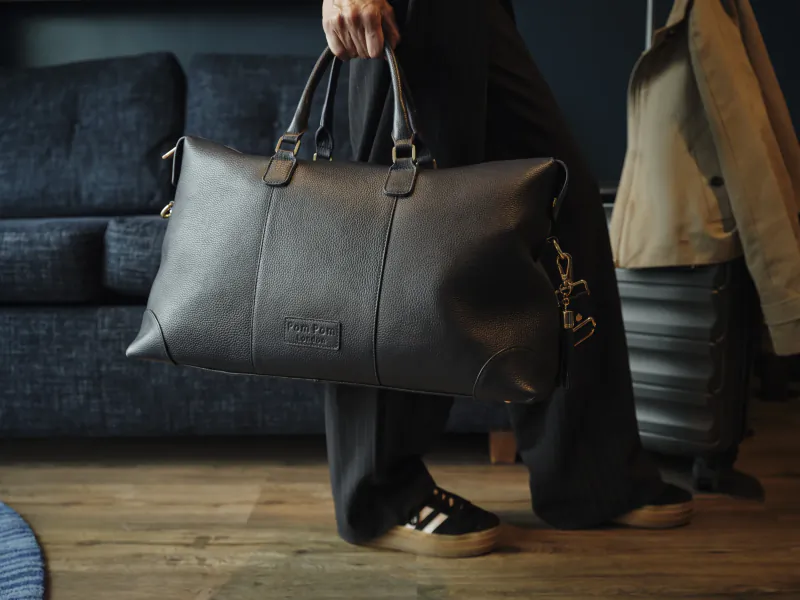 Person holding a large black leather duffel bag with gold hardware and Coach New York logo plate