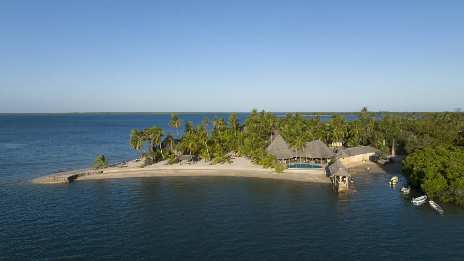 Aerial view of Manda Bay Lodge on a tropical island with thatched-roof buildings, palm trees, and a pool beside calm blue water