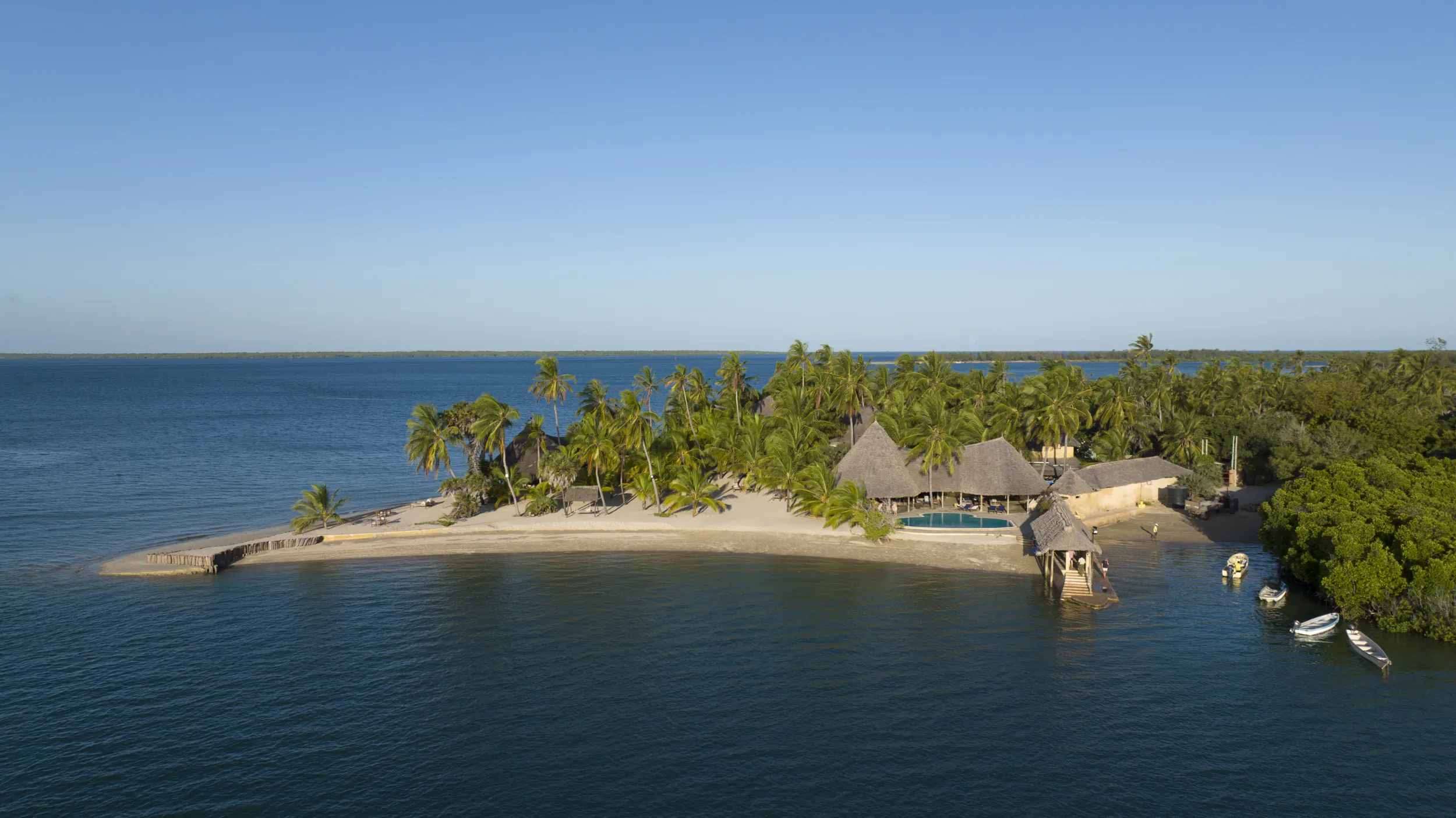 Aerial view of Manda Bay Lodge on a tropical island with thatched-roof buildings, palm trees, and a pool beside calm blue water
